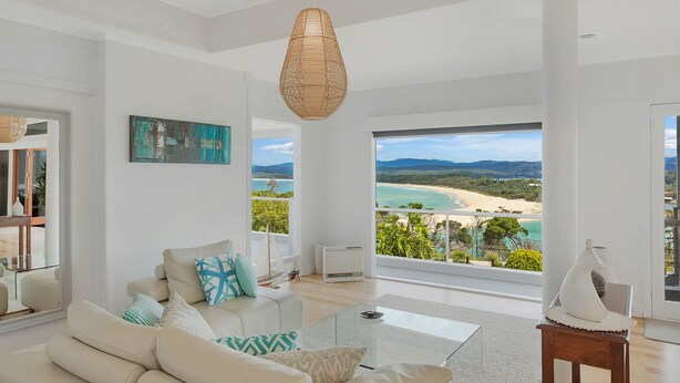 A spacious living room with a large window offering a view of a beach and mountains. - Lake 54 - Beautiful Views, Perfect Location! (Merimbula)
