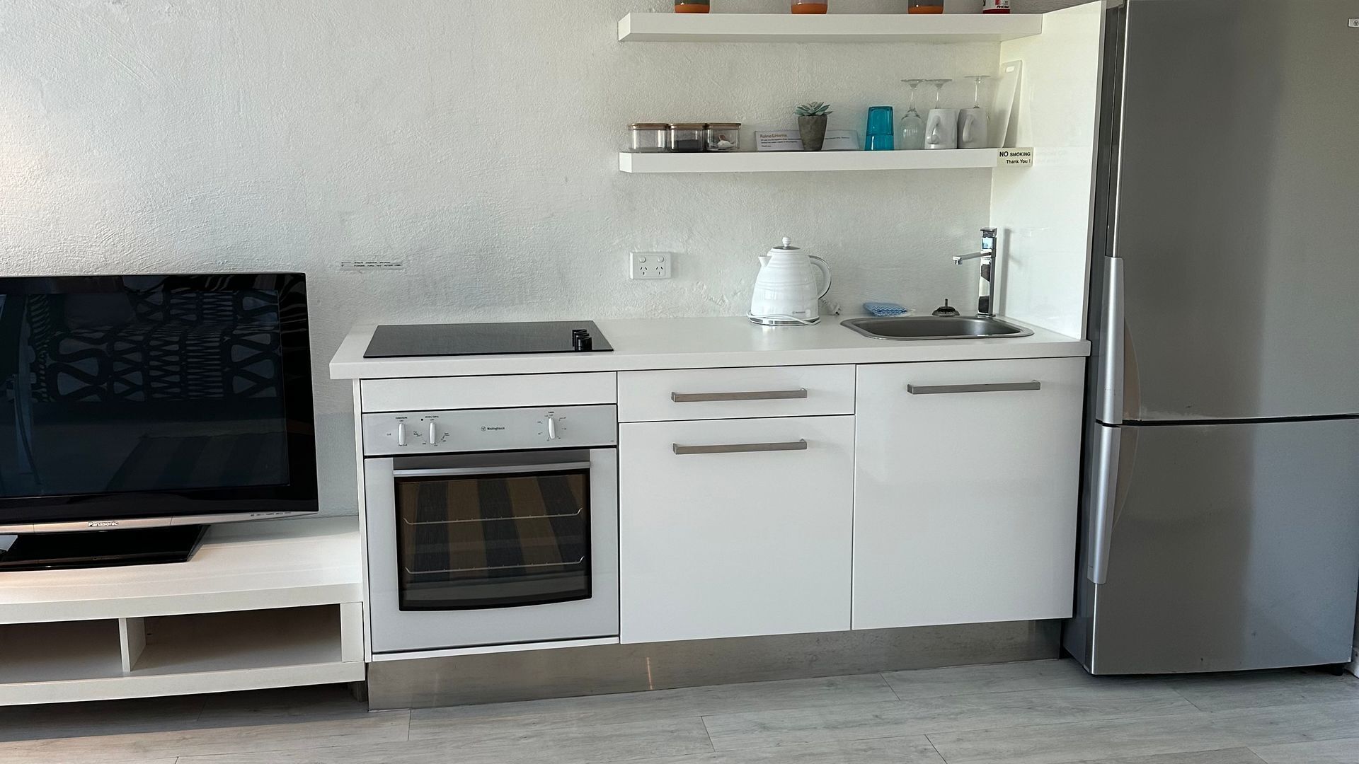A modern kitchen with white cabinetry, a built-in oven, a stainless steel refrigerator, and a flat-screen TV on a white stand.