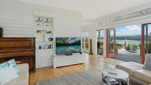 A modern living room with a large flat-screen TV, wooden furniture, and a view of a coastal landscape through large sliding doors. - Lake 54 - Beautiful Views, Perfect Location! (Merimbula)