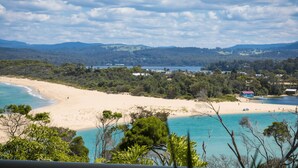 A coastal landscape with a sandy beach, clear turquoise waters, and a backdrop of hills and trees. - Lake 54 - Beautiful Views, Perfect Location! (Merimbula)