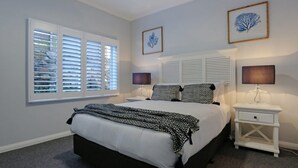 A bedroom with a large bed, two bedside tables, a window with blinds, and two framed wall art pieces.