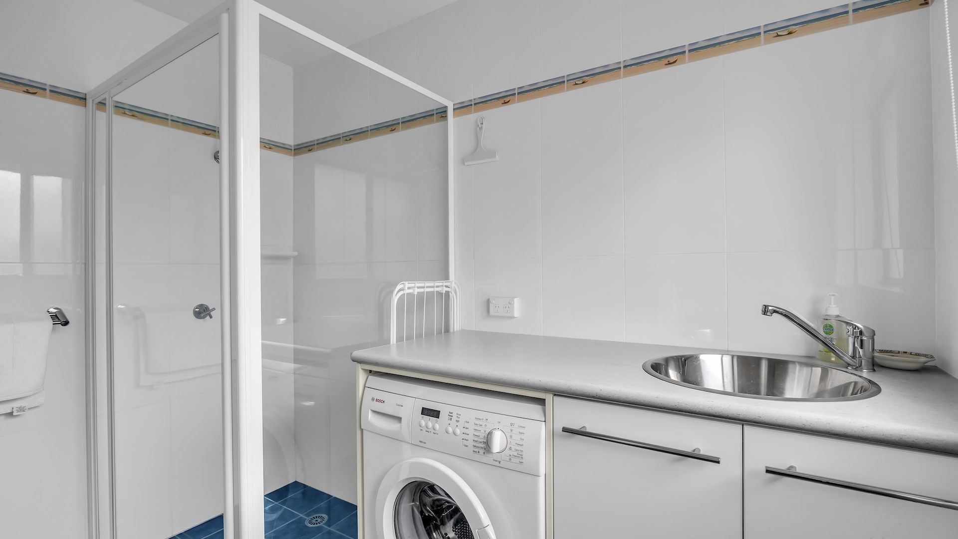 A modern bathroom with a glass shower enclosure, a built-in washing machine, and a stainless steel sink.