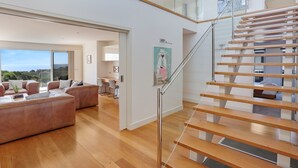 Un interior moderno con una escalera de madera, una sala de estar con sofá y una mesa de comedor, y vistas al exterior.