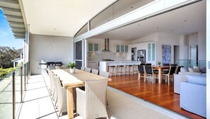 A modern kitchen with white cabinets and a stainless steel range hood, a dining area with a wooden table and wicker chairs, and a balcony with a grill and potted plant. - Short Point Quarters - Whole House (Merimbula)