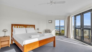 A modern bedroom with a wooden bed, a nightstand, a chair, and a large window offering a scenic view. - Our Beach House - with ocean views! (Merimbula)