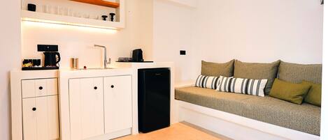 A compact kitchen with white cabinetry, a black refrigerator, and a coffee maker. There is a sofa with cushions in the adjacent living area.