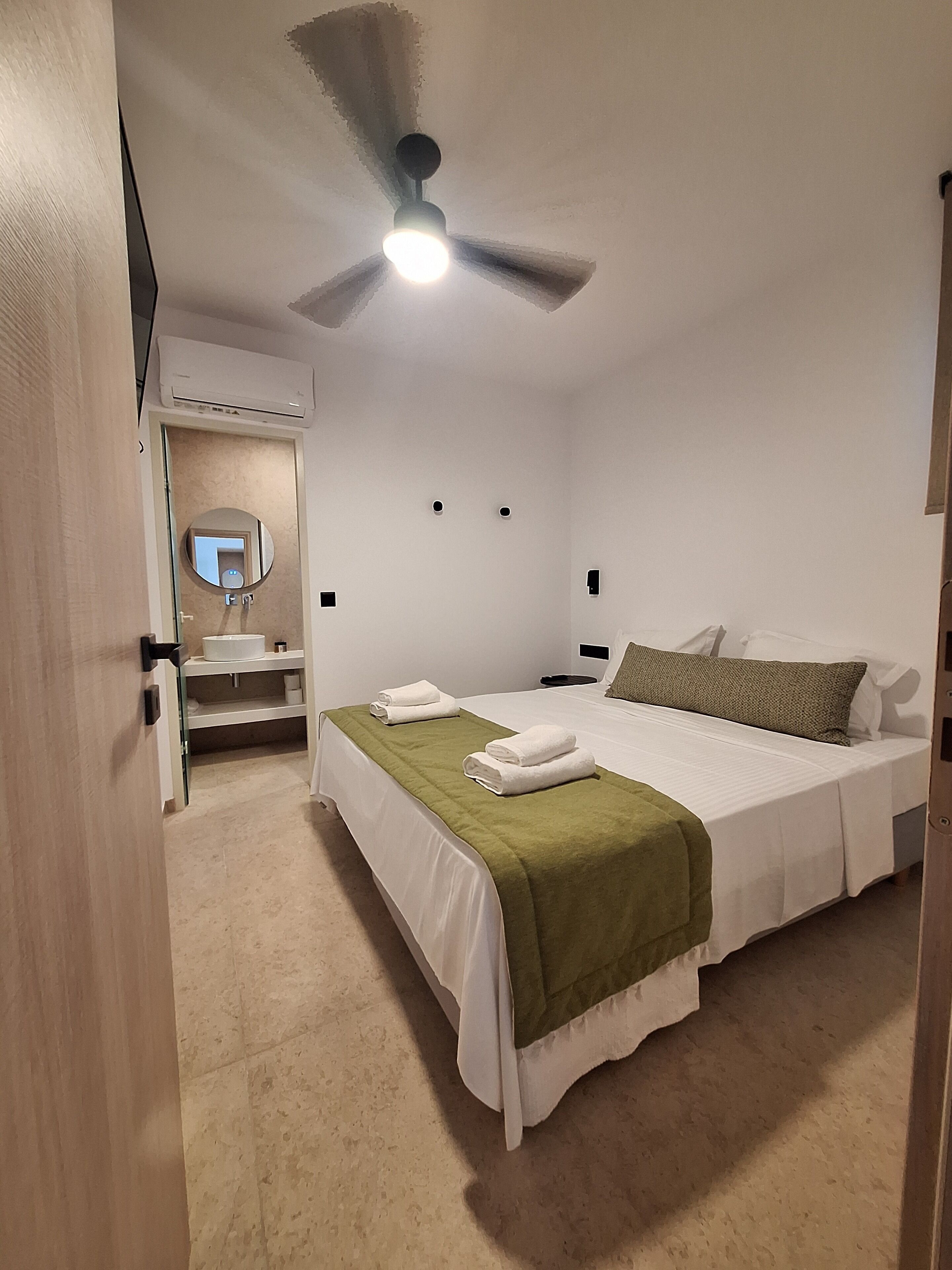 A hotel room with a bed, a green throw blanket, white towels, a ceiling fan, a mirror, and a small bathroom with a sink and toilet.