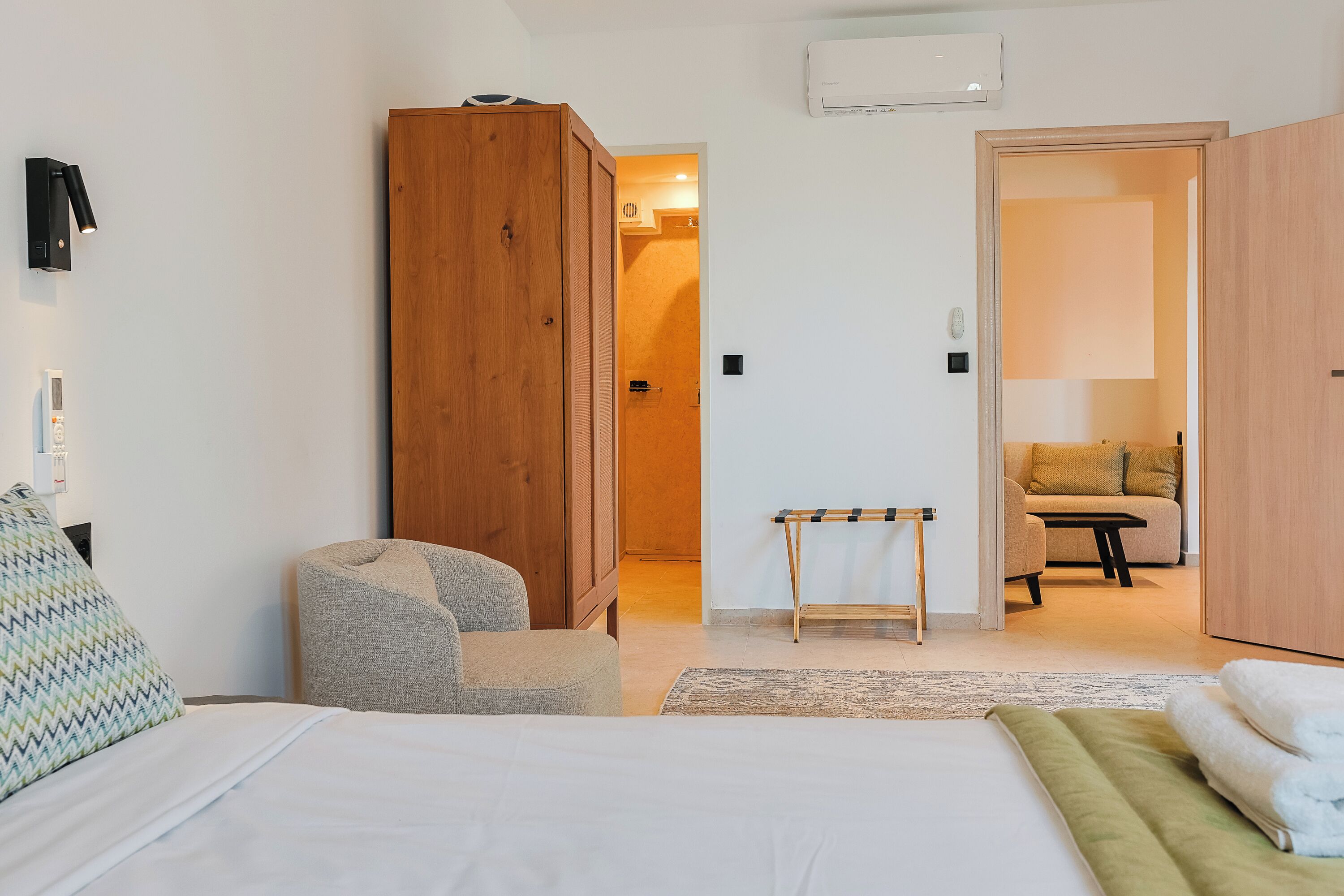 A hotel room with a bed, a wooden wardrobe, a small table, a chair, and a sofa in the background.