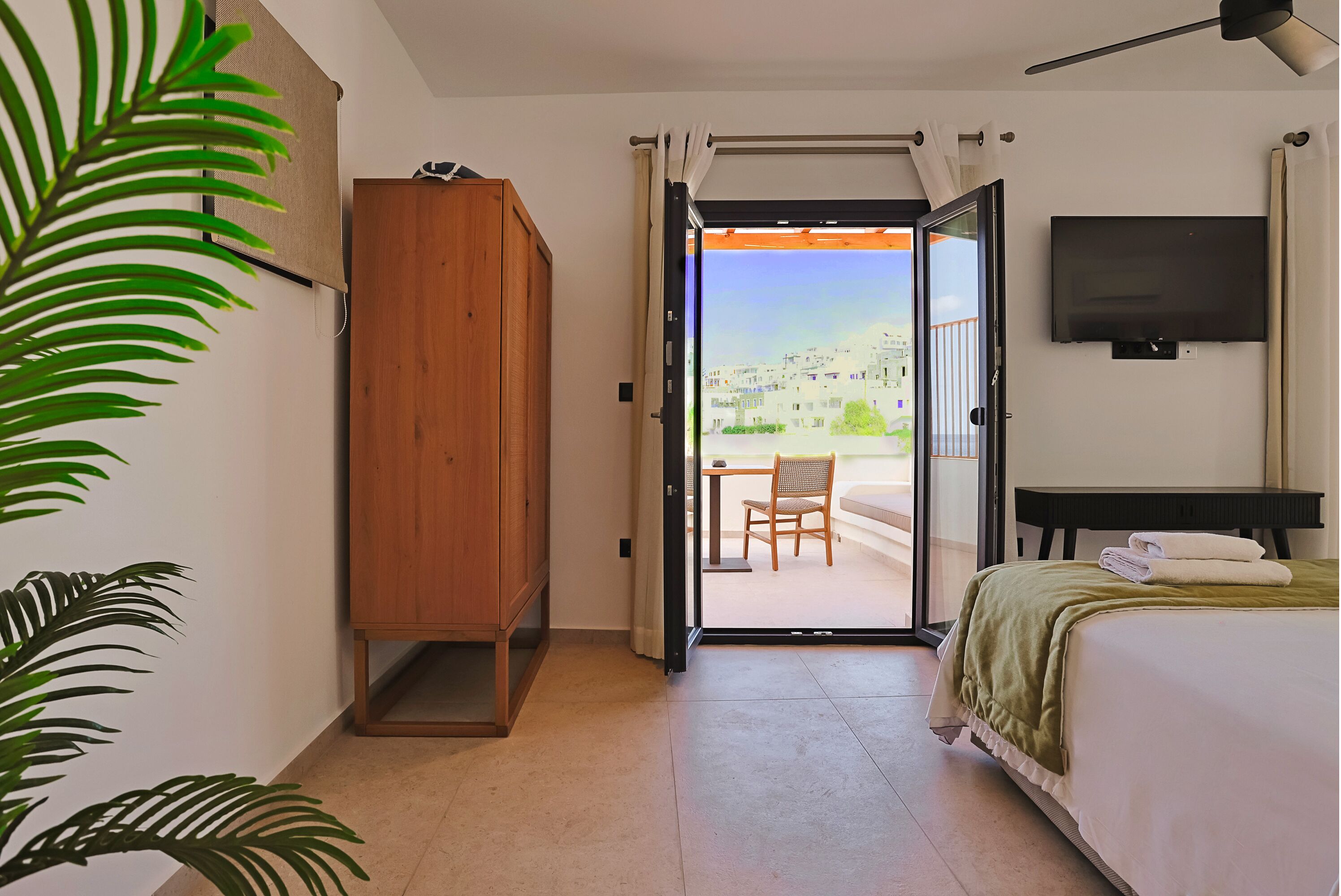 Habitación de hotel con cama, armario de madera, televisión y puerta corredera que da a un espacio exterior con mesa y sillas.