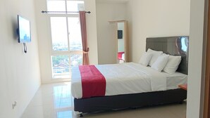 A bedroom with a bed, a TV mounted on the wall, and a window with curtains. - PLUM Dreamscape Bale Hinggil Surabaya (Surabaya)