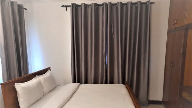 A single bed with a wooden frame and white bedding in a room with grey curtains and a wooden wardrobe. - Spacious Family Home  (Arusha)