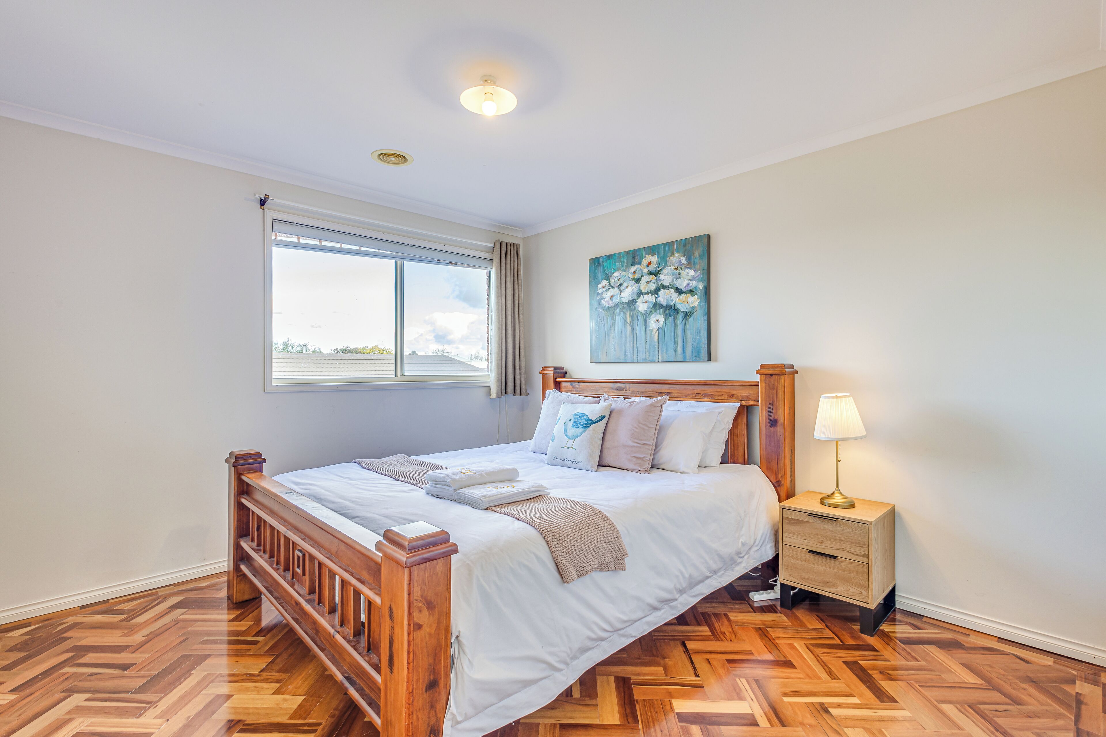 A bedroom with a wooden bed, a nightstand with a lamp, a window with curtains, and a painting on the wall.