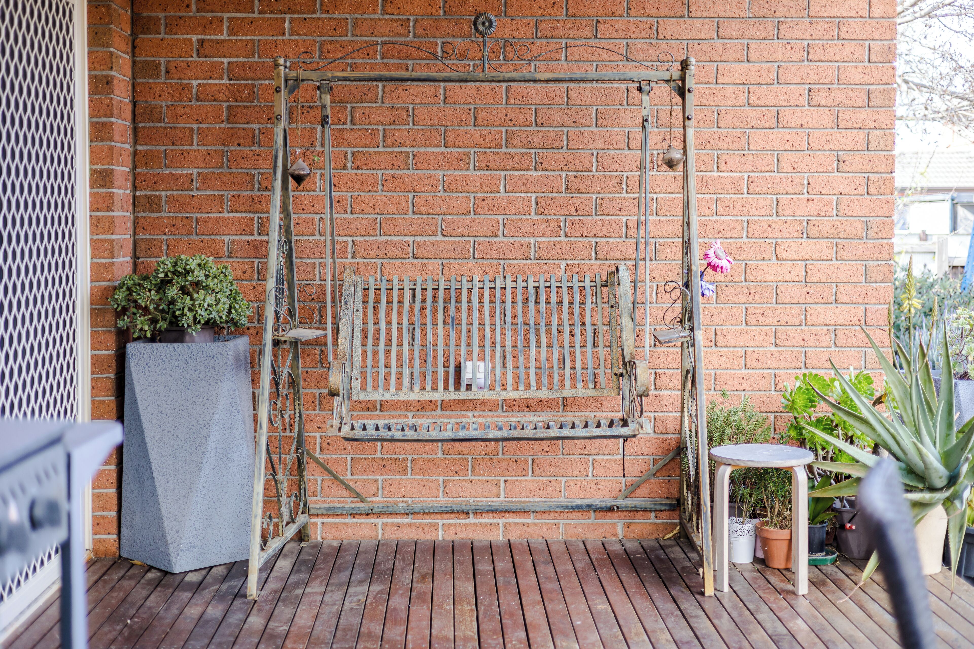 Un columpio metálico frente a un muro de ladrillo, una planta en maceta y una pequeña mesa con más plantas en macetas sobre una terraza de madera.