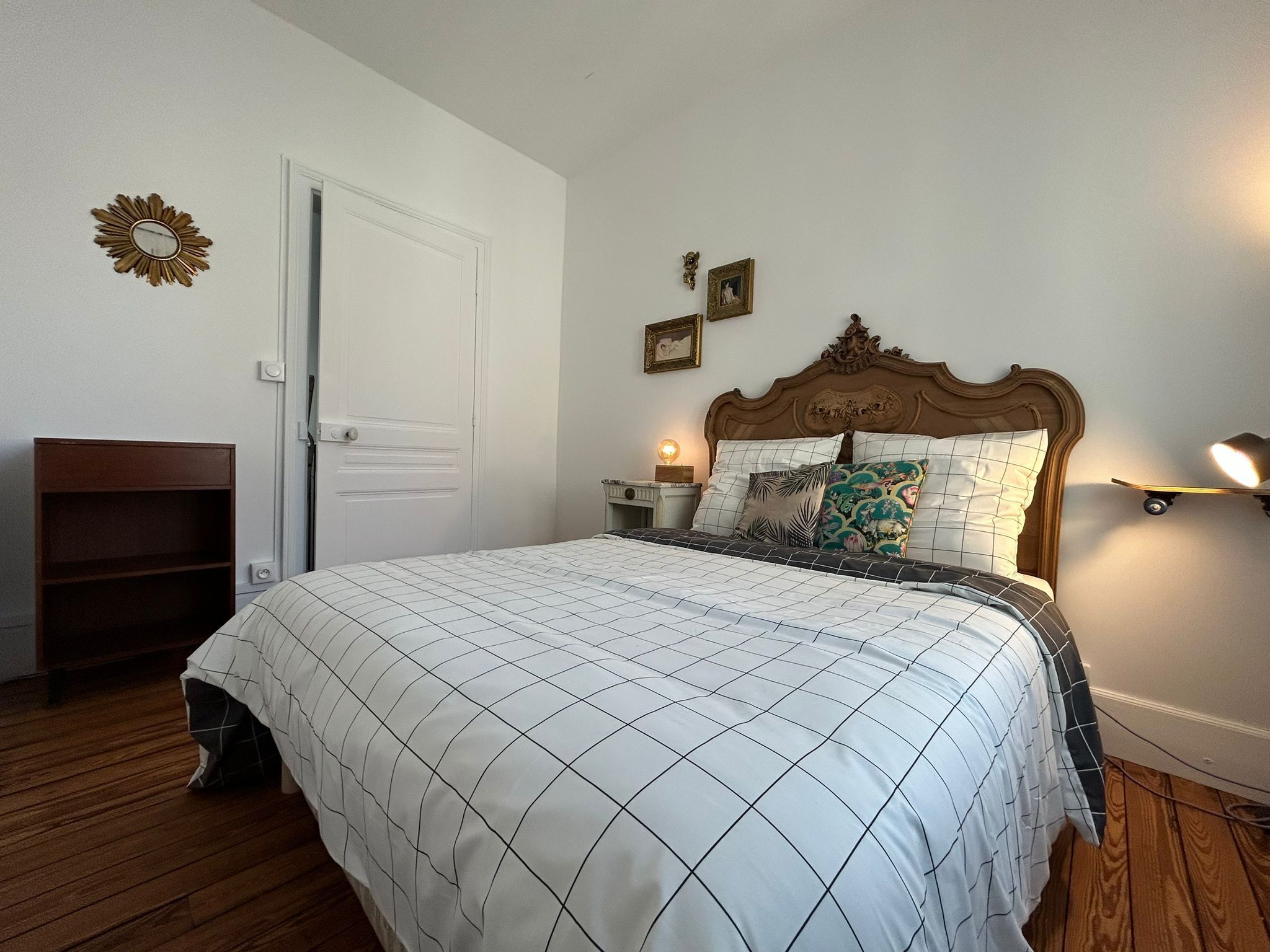 Quarto com cama grande, cabeceira de madeira, mesa de cabeceira com abajur, prateleira pequena e uma porta.