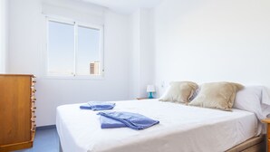 A bedroom with a bed, a wooden dresser, a nightstand, a lamp, and a window with a view of buildings. - Mar de Oropesa II by Interhome (Oropesa del Mar)