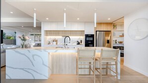 A modern kitchen with a marble island, two wicker chairs, pendant lighting, and a stainless steel refrigerator.