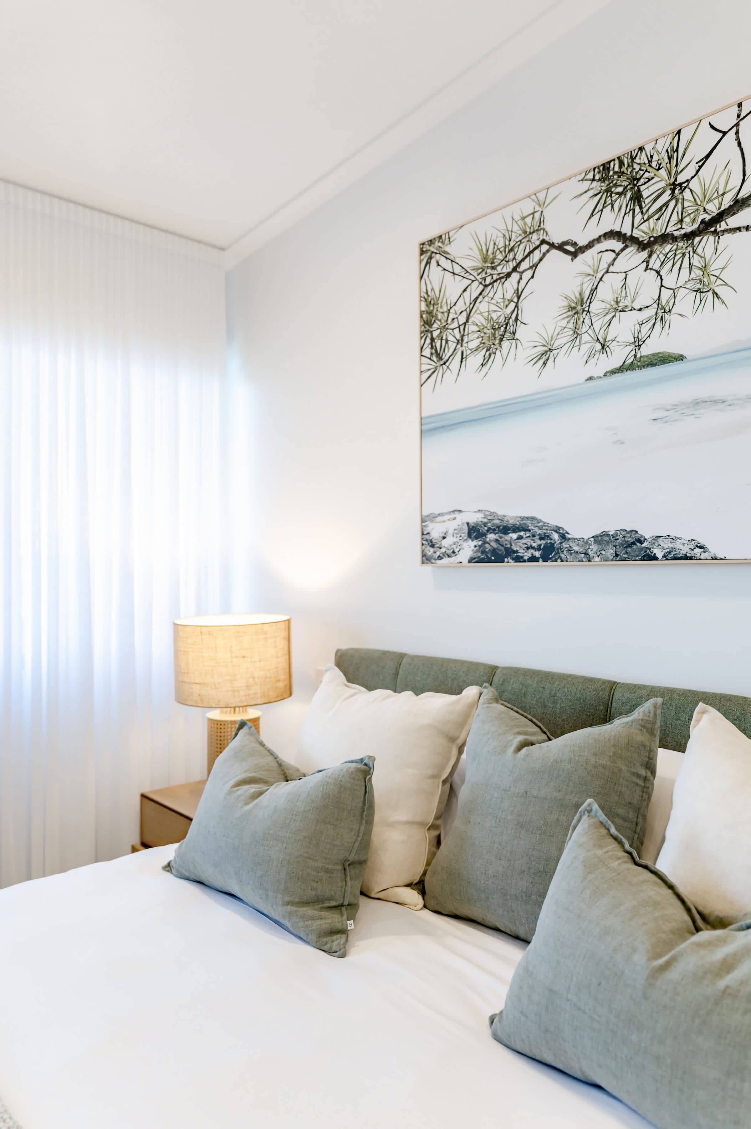 A bedroom with a bed, pillows, a nightstand, a lamp, and a wall-mounted painting of a beach scene.