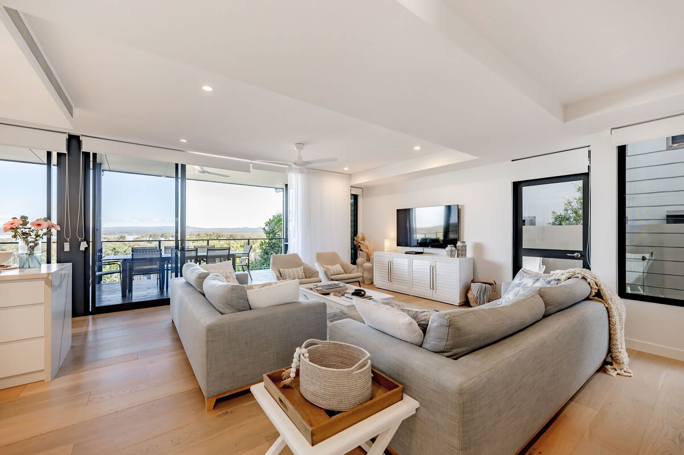 A modern living room with a sofa, coffee table, and TV. There is a balcony with outdoor furniture.