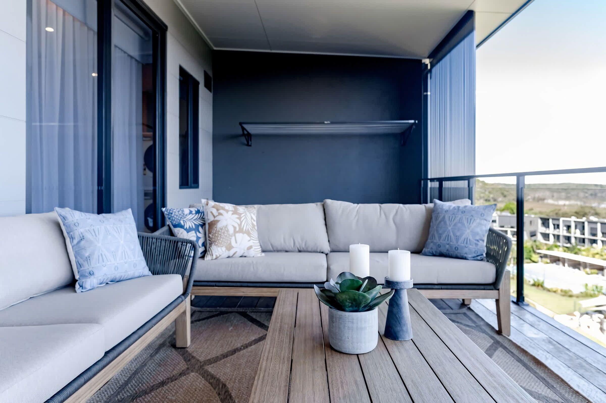 A modern outdoor seating area with a sofa and cushions, a wooden table with candles, and a view of the landscape.