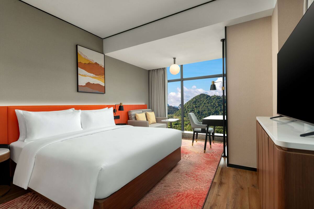 Una camera d'albergo con un letto grande, una scrivania, una sedia, una TV e una finestra vista montagna.