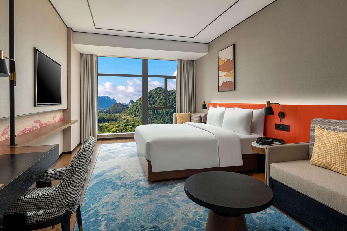 Habitación de hotel con una cama grande, un escritorio, una silla, un sofá, un televisor y una ventana con vista a la montaña.