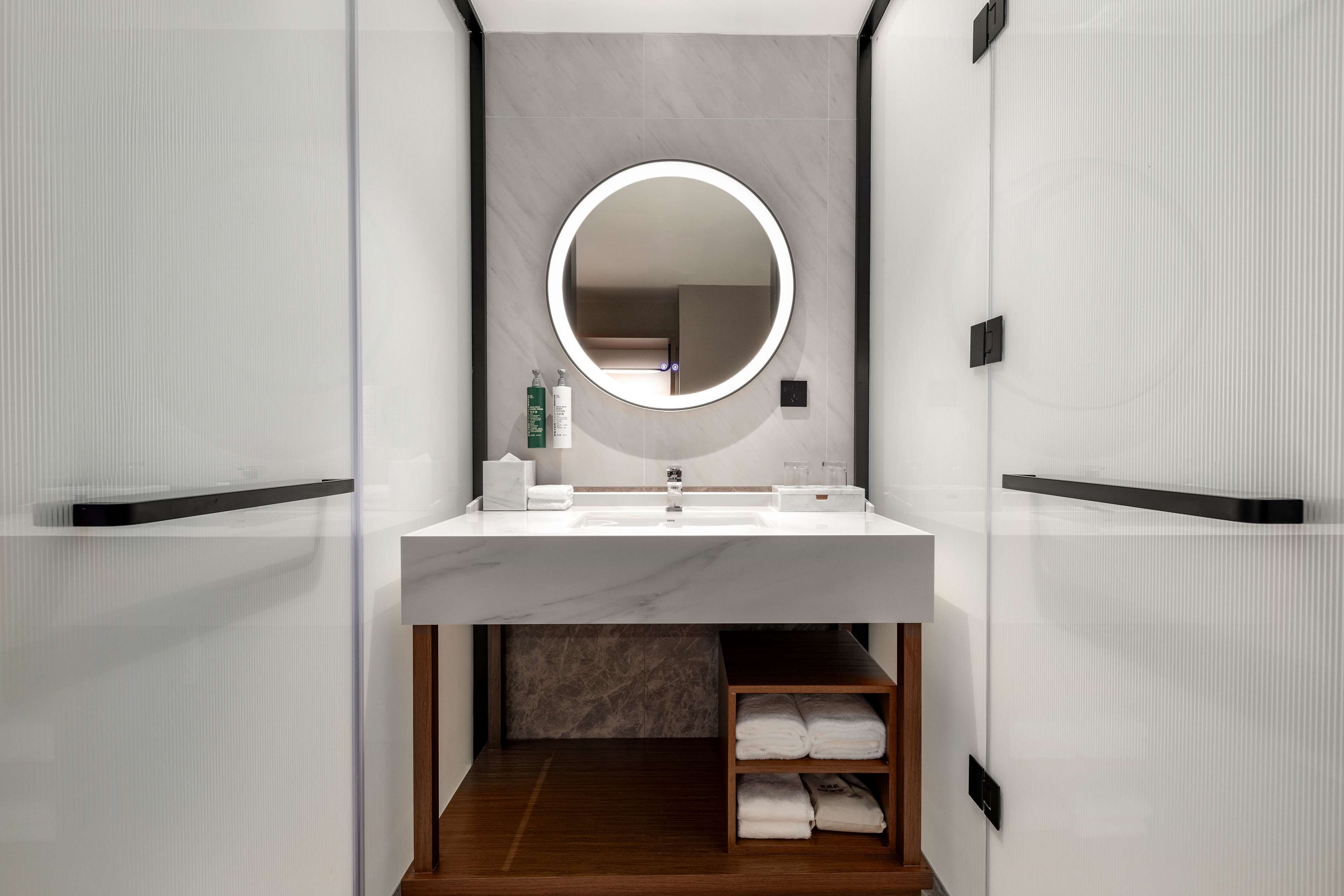 A modern bathroom with a round mirror, white sink, and wooden shelf with towels.