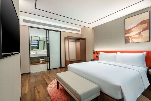 Une chambre d’hôtel moderne dotée d’un grand lit, d’une banquette, d’un bureau, d’une télévision et d’une salle de bain visible à travers une porte en verre.
