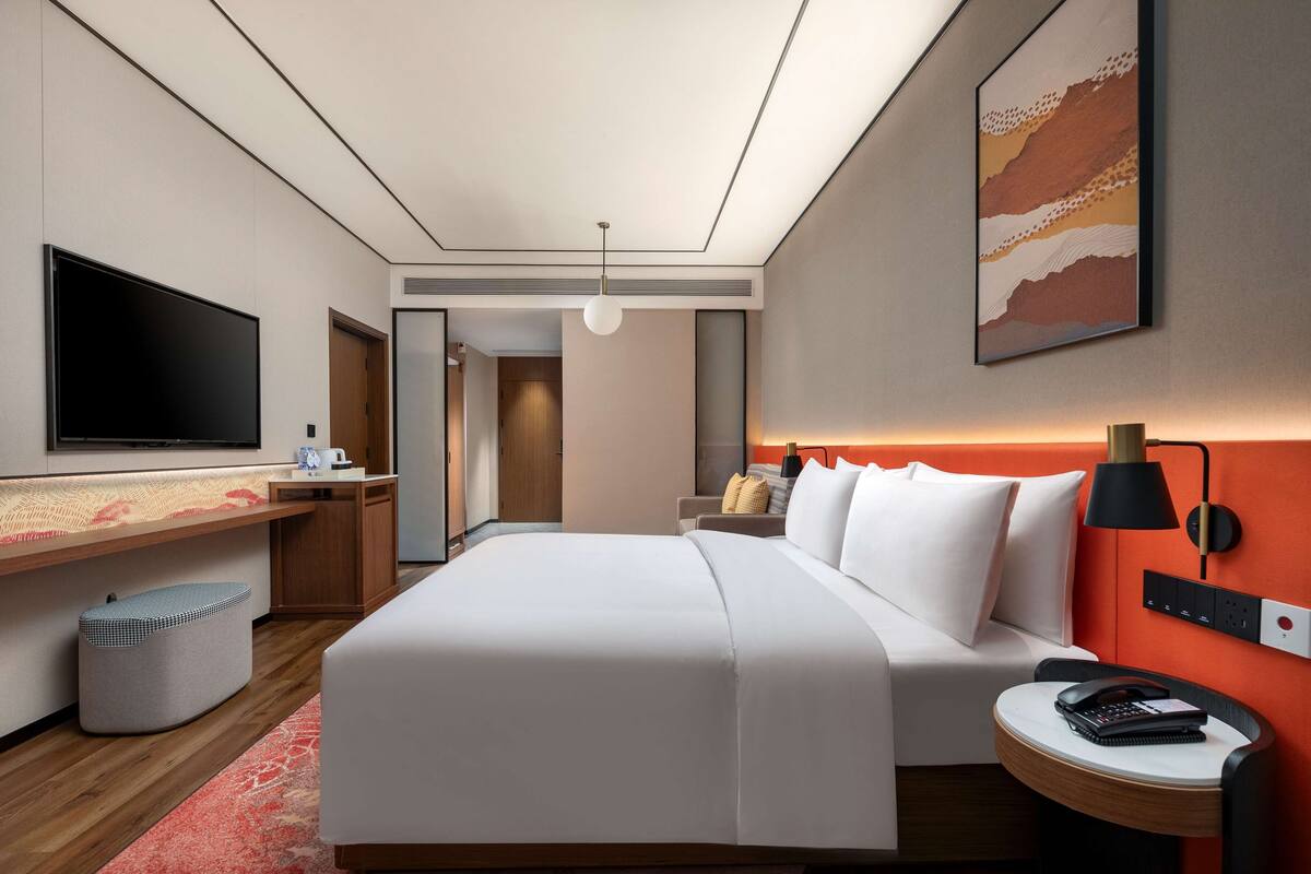 Una camera d'albergo moderna con un letto grande, una TV a schermo piatto, una scrivania con una sedia, un cestino e una lampada a parete.