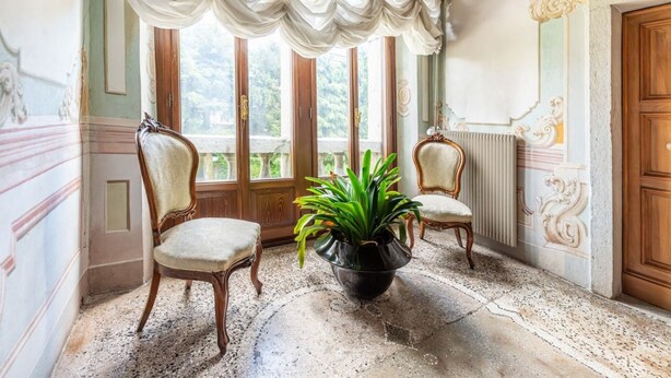 Ein Zimmer mit zwei Stühlen, einer Topfpflanze, einem großen Fenster mit Vorhängen und aufwendigen Wandverzierungen.