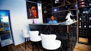 A bar area with a counter, white chairs, a drink dispenser, and a wall with a framed picture and shelves of bottles. - Vic & J Lounge and Hotel (Asaba)