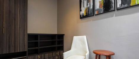 A modern interior with a white armchair, a wooden stool, a dark wood cabinet, and abstract art on the wall.