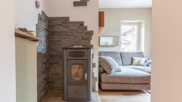 A living room with a stone fireplace, a sofa, and a window. - Appartamento Casa Tre Colli (Trentino-Alto Adige)