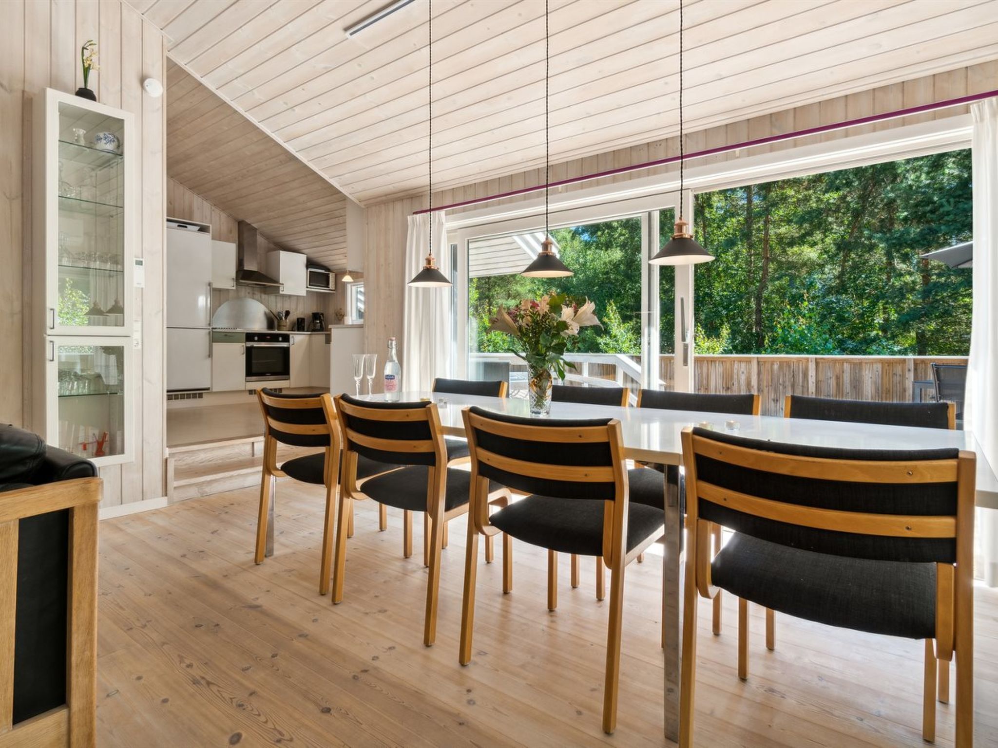 A modern kitchen with white cabinets, a dining area with a wooden table and chairs, and large windows offering a view of the greenery outside.