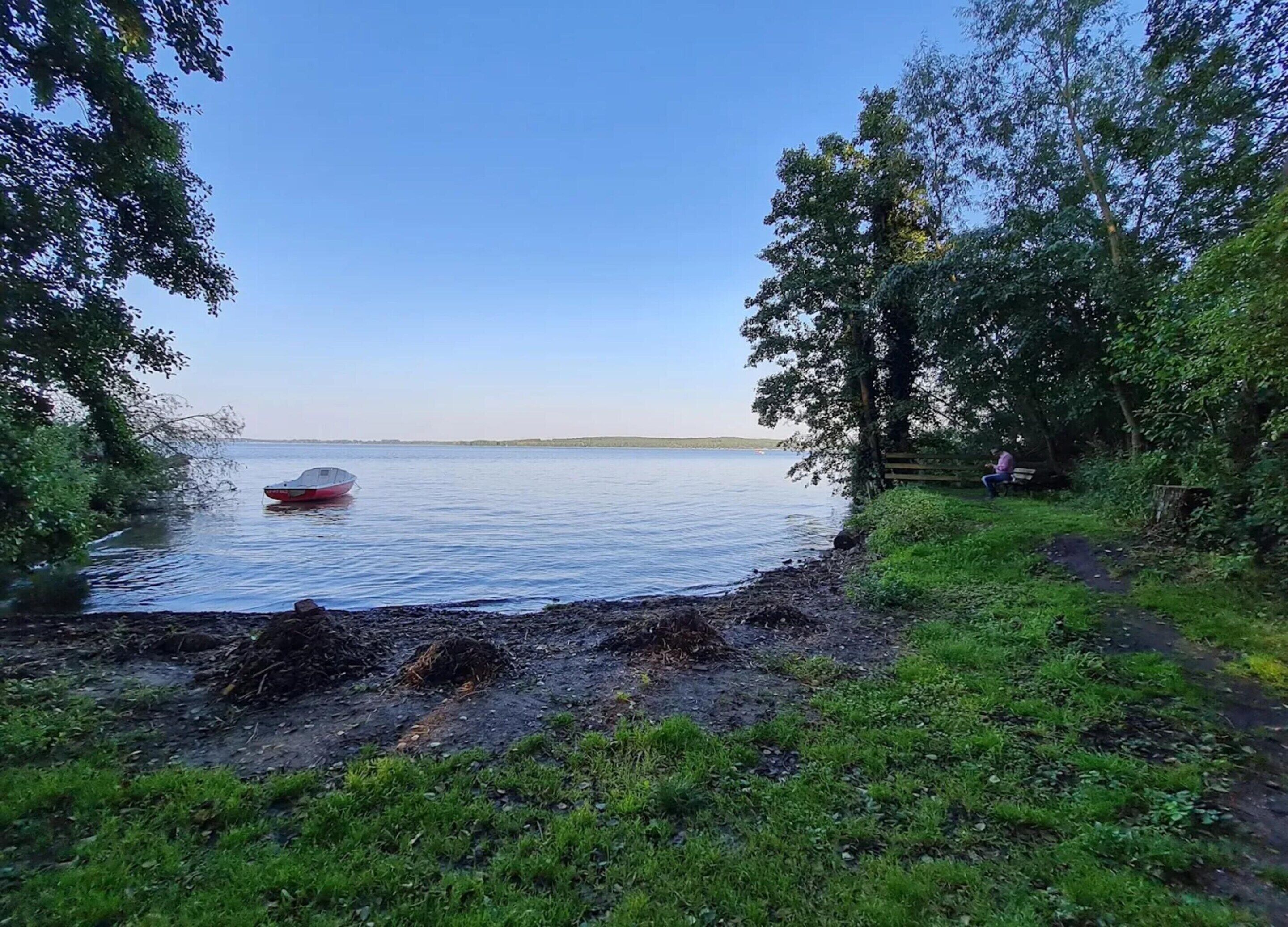 Eine friedliche Seenlandschaft mit einem roten Boot auf dem Wasser, umgeben von üppigem Grün und blauem Himmel.