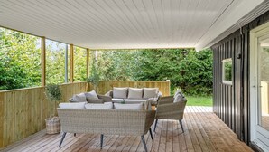 A covered outdoor area with a wooden deck, wicker furniture, and a view of a lush garden.