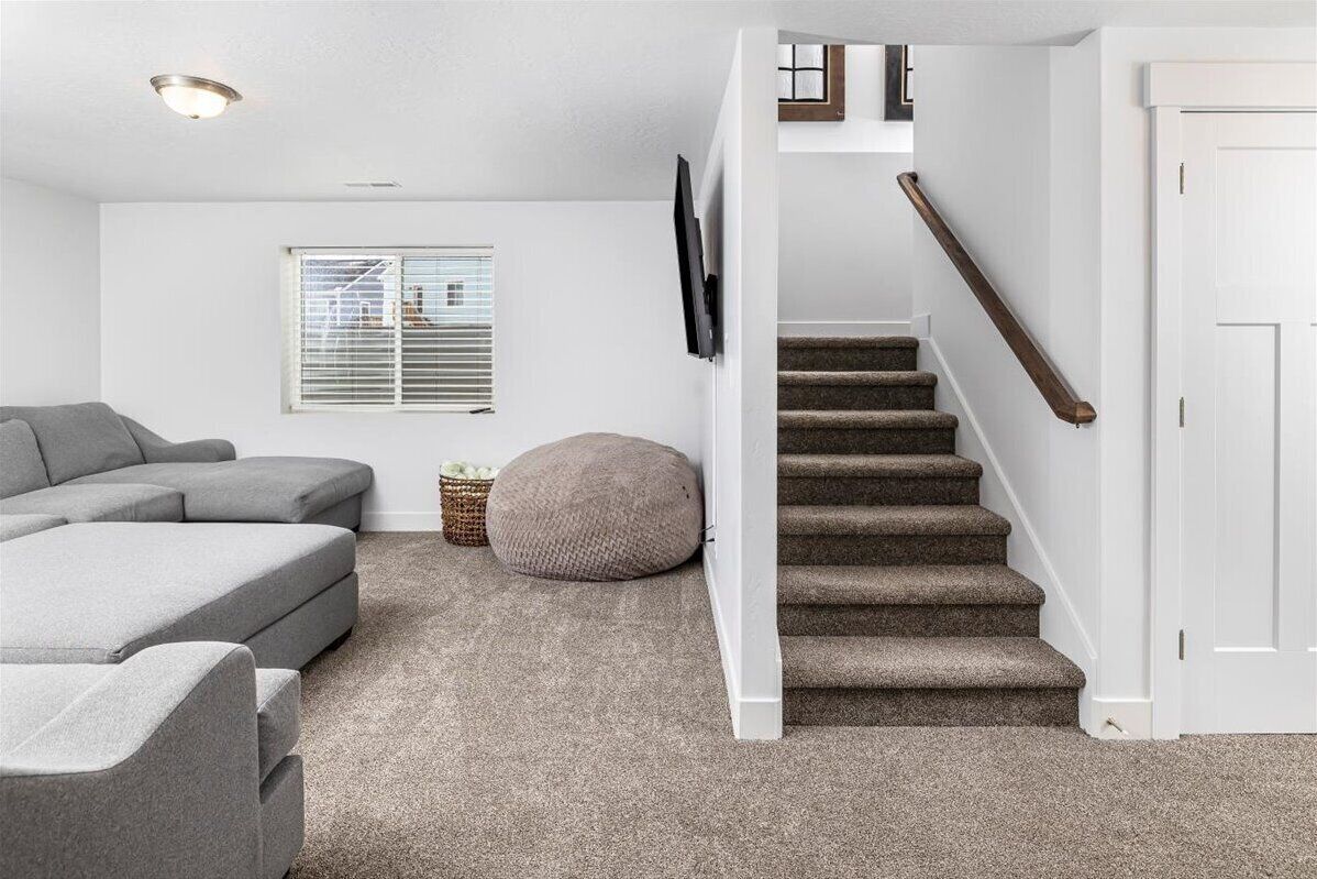 Un salon avec un canapé d’angle gris, un pouf rond, un escalier doté d’une rampe en bois et une porte blanche.