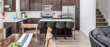 A modern kitchen with dark wood cabinets, stainless steel appliances, a central island with bar stools, and a dining area with a wooden table and chairs.