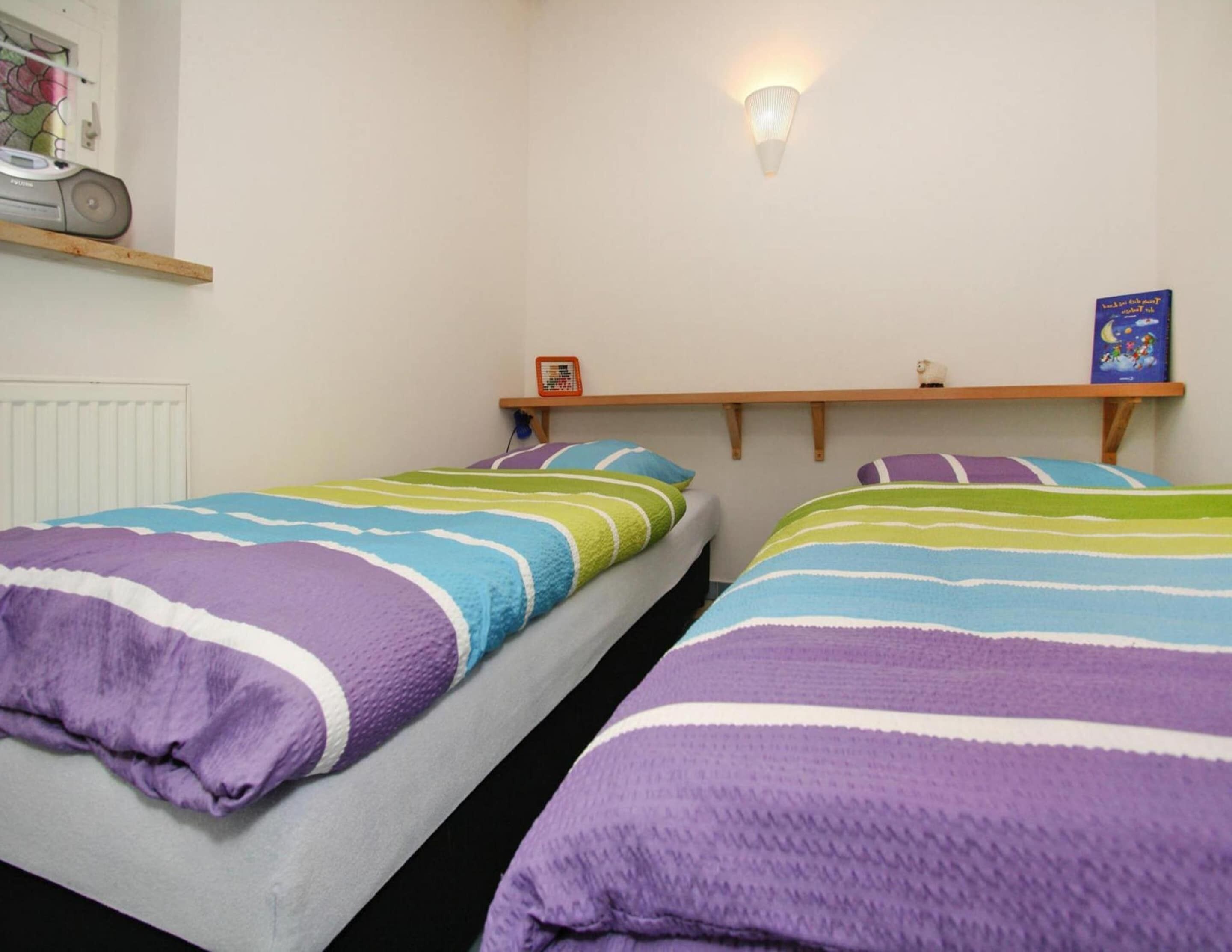 Une chambre avec deux lits, chacun doté de draps à rayures, d’un luminaire fixé au mur et d’une étagère sur laquelle se trouvent un livre et un petit objet.