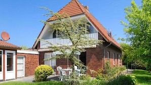 Een huis met twee verdiepingen, voorzien van een rood pannendak, een schotelantenne en een kleine tuin met een witte stoel en tafel.