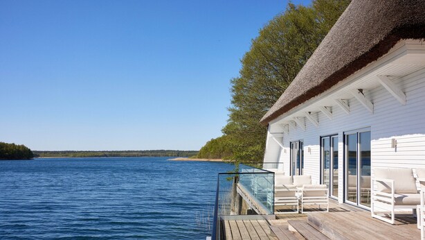 A white building with a thatched roof, a wooden deck, and a glass railing overlooking a body of water. - Doppelhaushalften im Naturresort Drewitz (Nossentiner Huette)