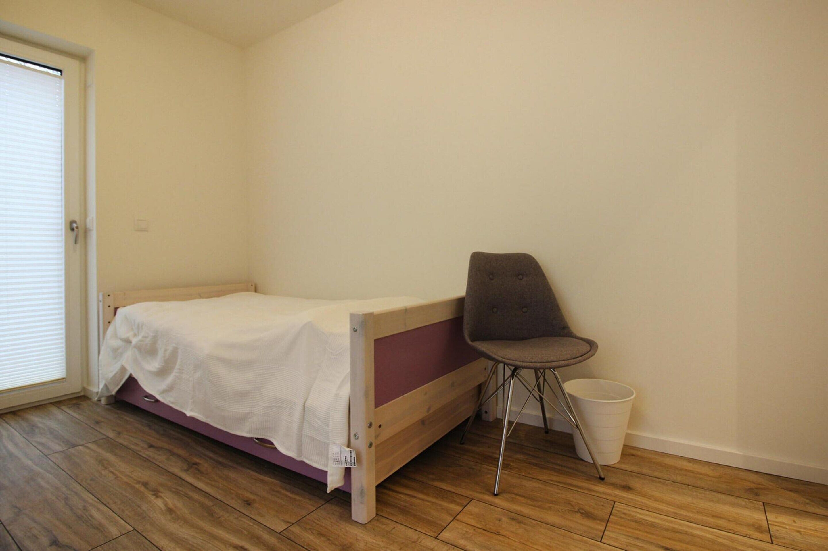 A small, single-bed room with a wooden frame, a grey chair, and a white bucket.