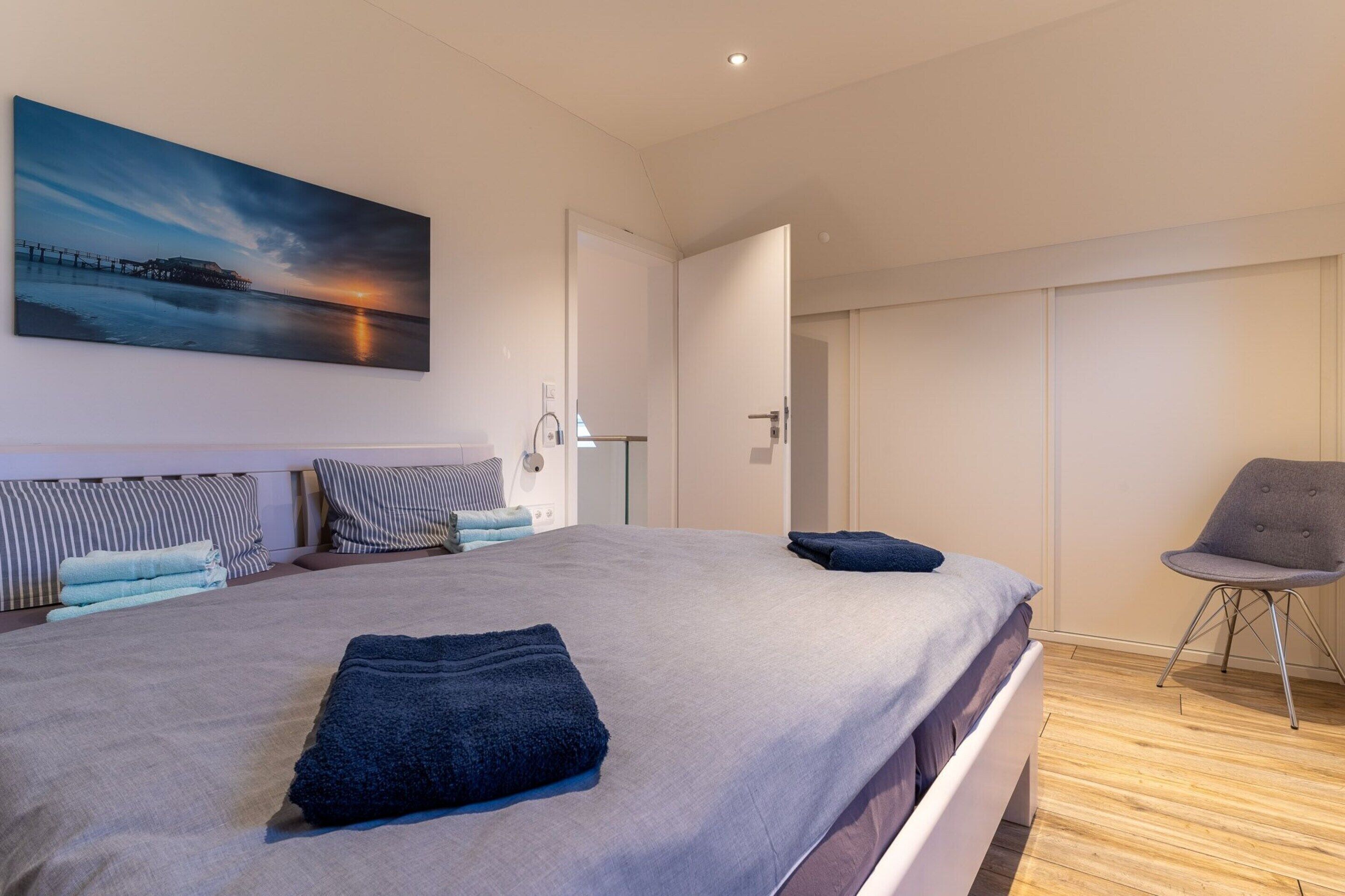 A modern hotel room with a large bed, a chair, a painting, and a view of the sea.