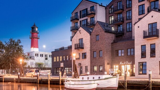 Un hotel sul lungomare con edifici in mattoni, una barca ormeggiata in primo piano e un faro sullo sfondo.