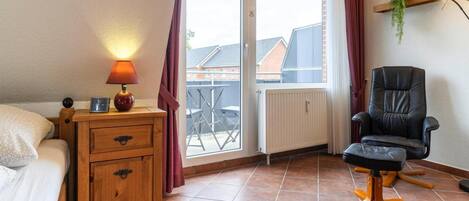 Ett sovrum med en säng, ett träbord som står vid sängen, en lampa, en stol, ett element och ett fönster med gardiner.