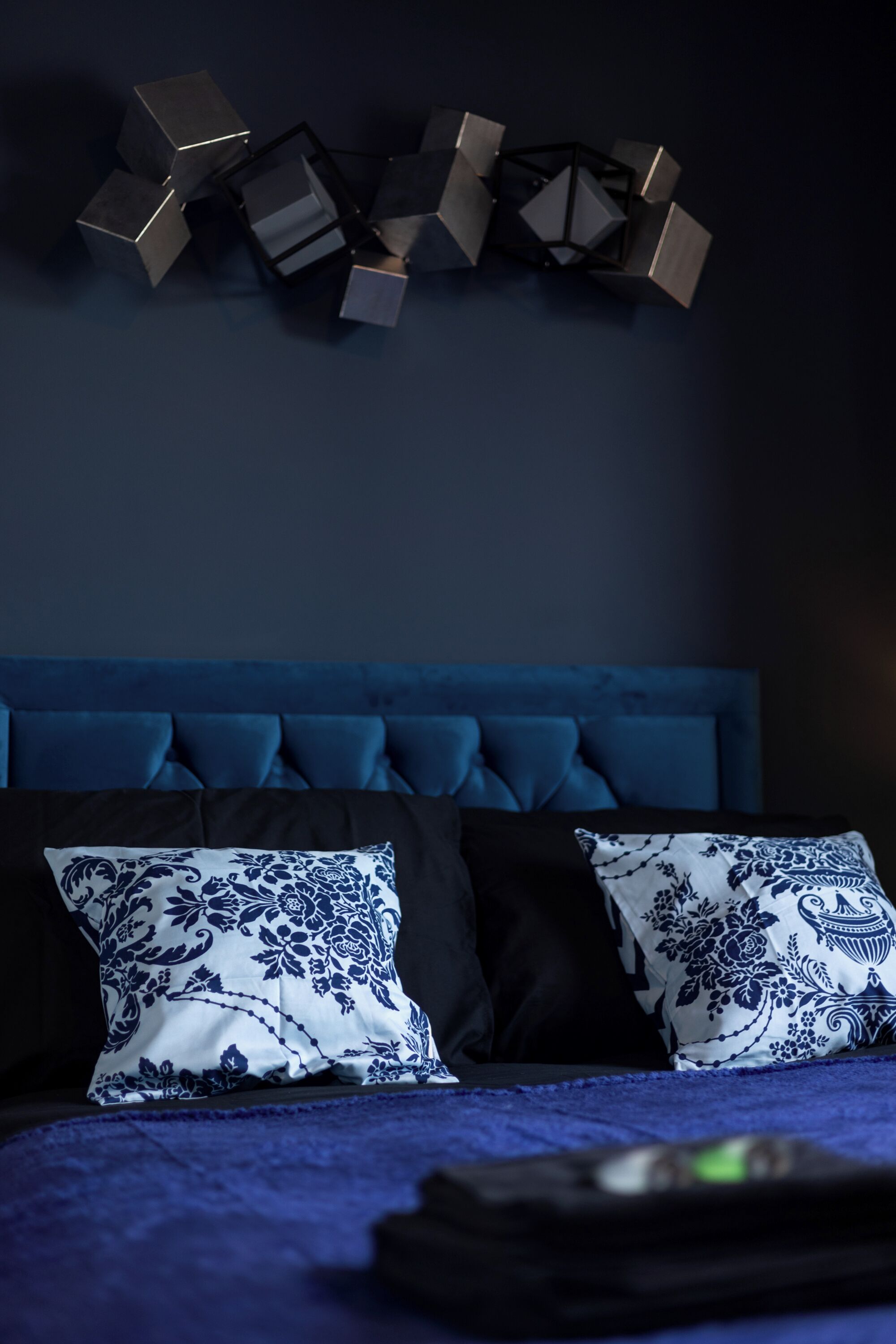 A bedroom with a blue headboard, two patterned pillows, and a geometric wall decoration.