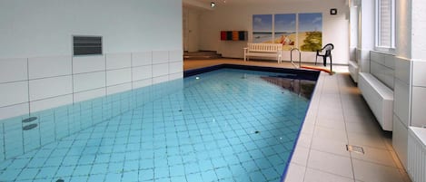 An indoor swimming pool with a tiled floor, a bench, and a view of the sea through the windows.