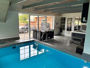 Une piscine intérieure avec vue sur l’extérieur, un espace détente avec des fauteuils et un barbecue.