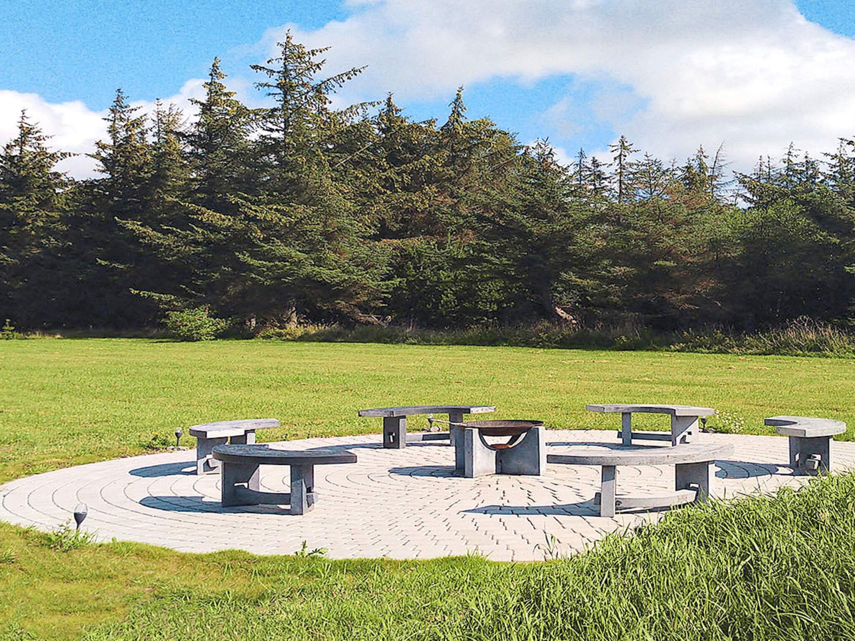 A circular outdoor seating area with benches and tables, surrounded by a grassy field and dense trees.