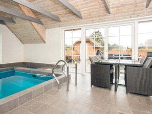 A swimming pool with a glass enclosure, a dining area with wicker furniture, and a view of a brick building through large windows.