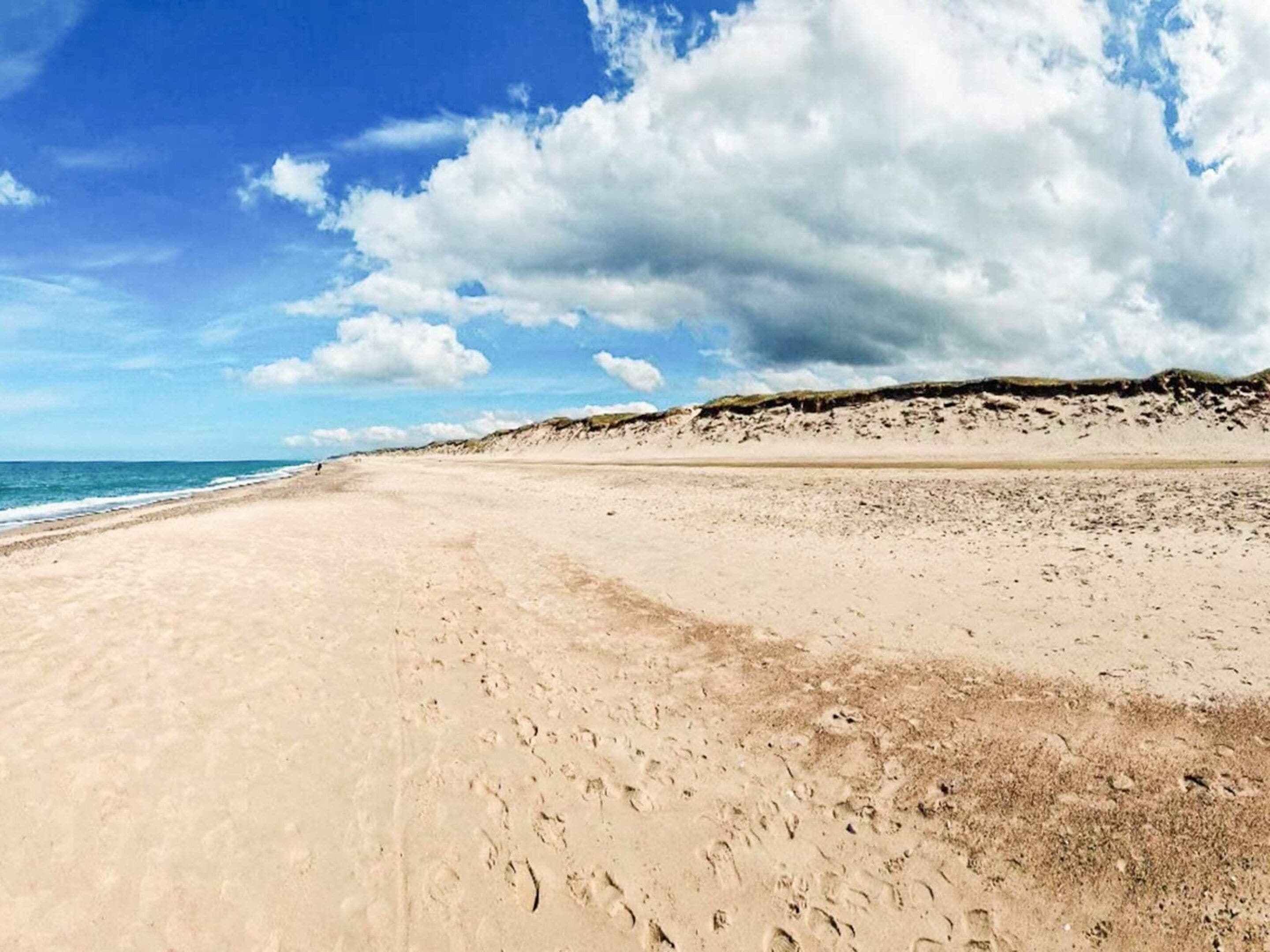 A wide sandy beach with footprints leading to the water, a clear sky, and a distant dune.